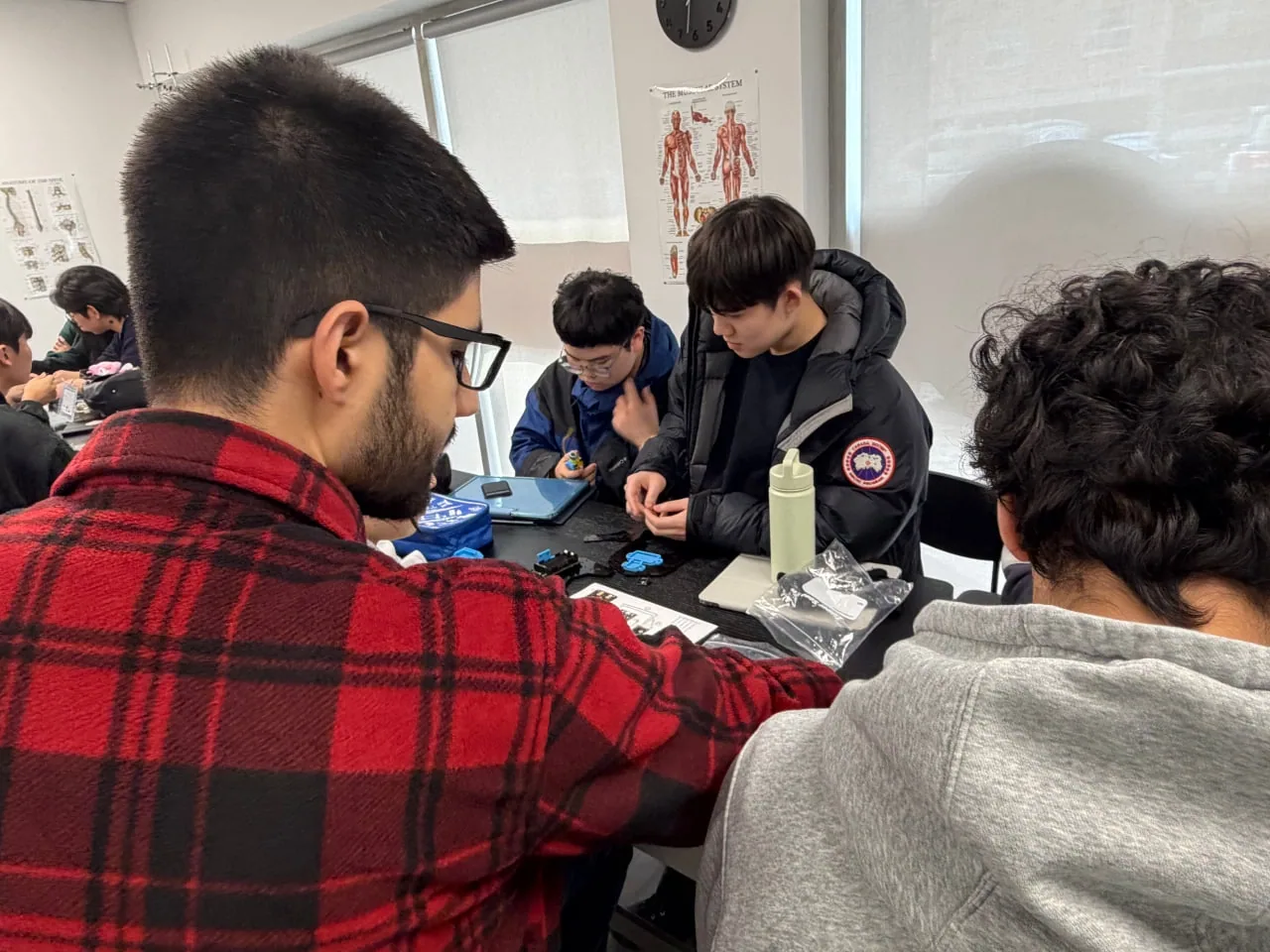 Instructor in a plaid shirt working alongside teens assembling kits at a classroom table