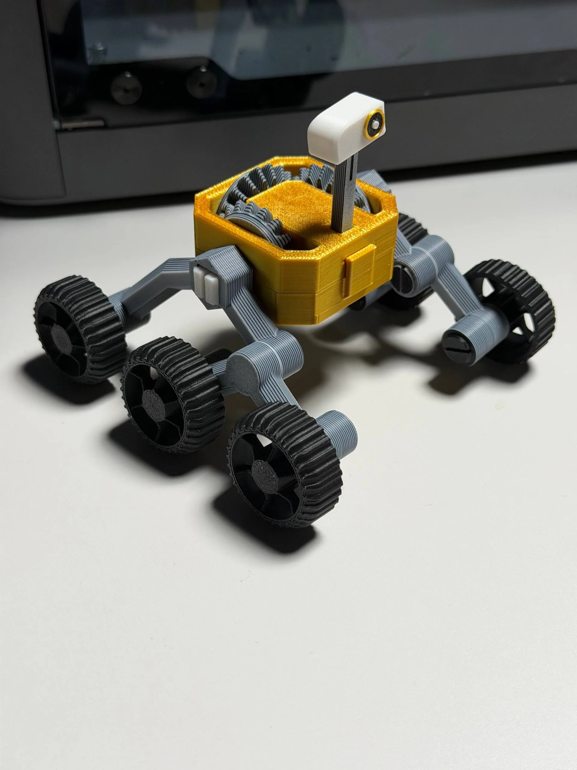 3D-printed six-wheeled rover model with a yellow body and camera head on a table
