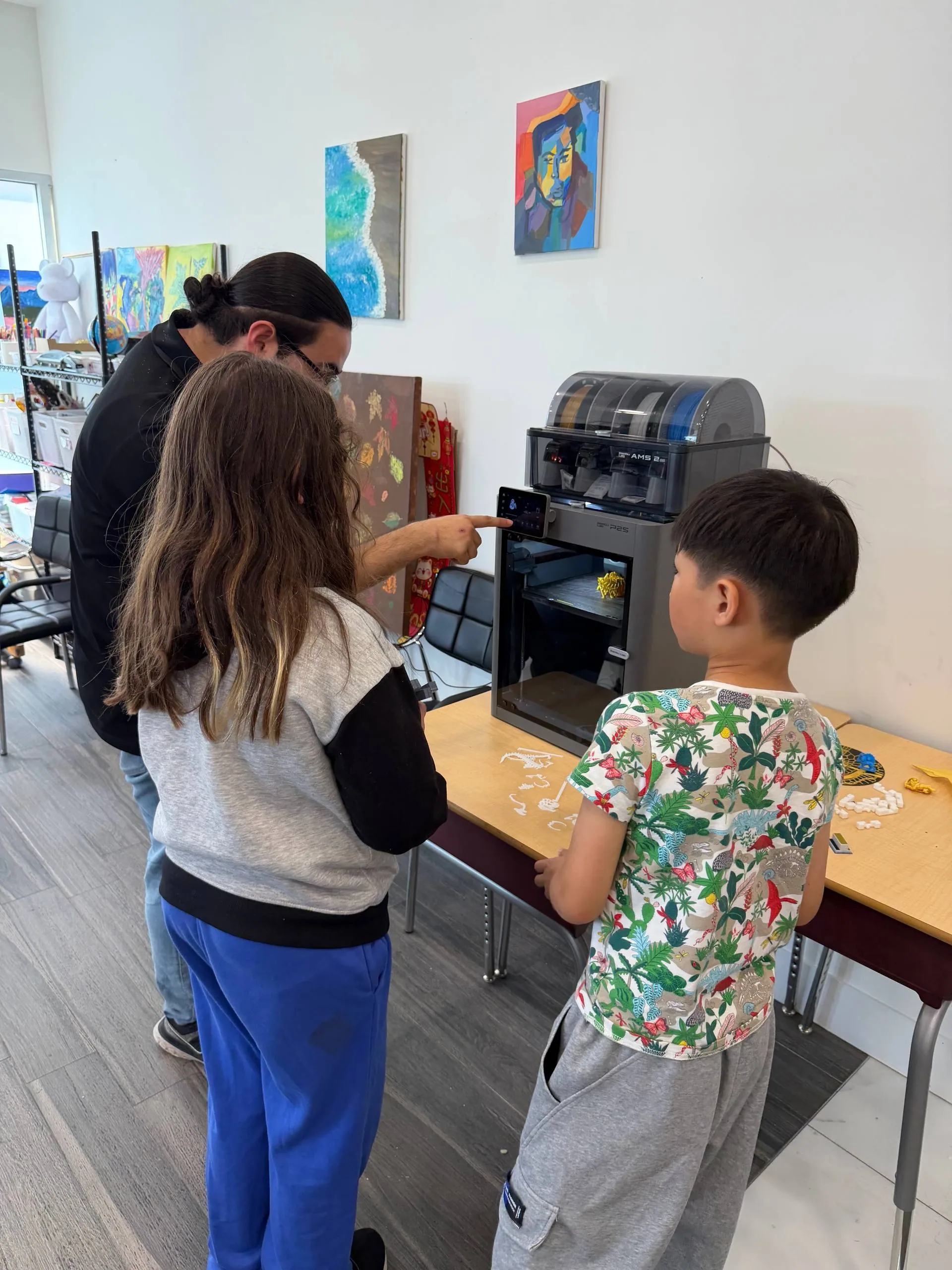 Instructor pointing at a 3D printer's touchscreen as two children look on